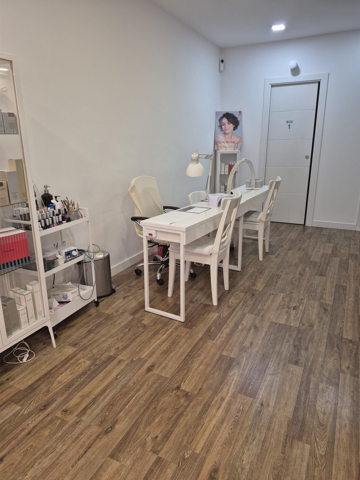 Un luminoso y minimalista rincón de manicura con dos escritorios blancos, sillas a juego y un carrito auxiliar sobre un suelo que imita la madera.