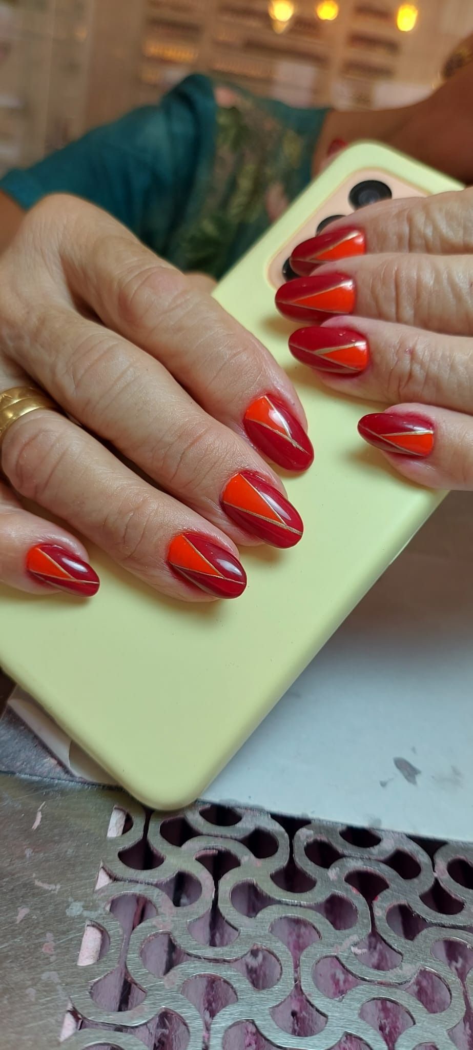 Manos que sostienen un teléfono verde claro, mostrando uñas bien cuidadas en forma de almendra con un diseño diagonal bicolor en rojo y naranja.