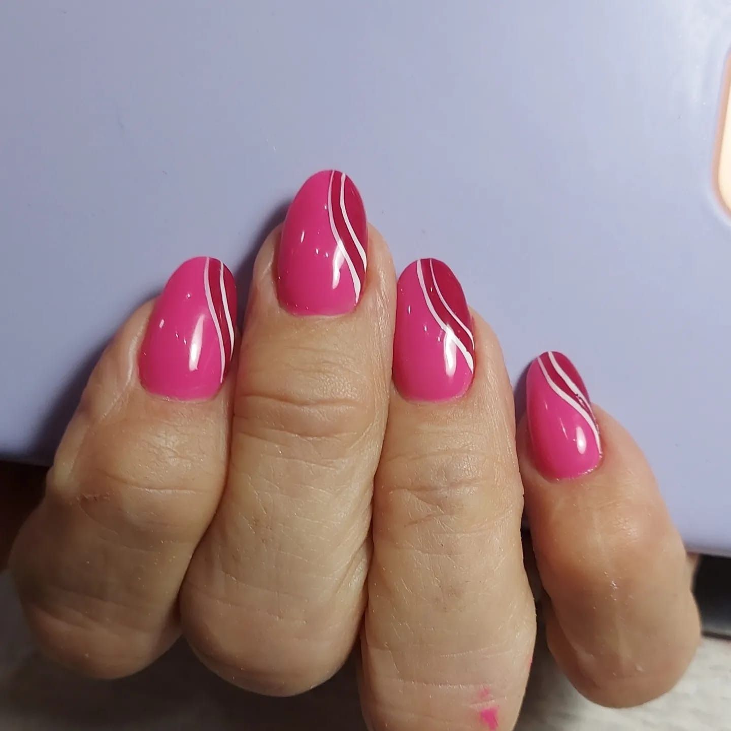 Una mano con uñas almendradas pintadas de color rosa brillante, cada una adornada con dos finas líneas blancas onduladas.