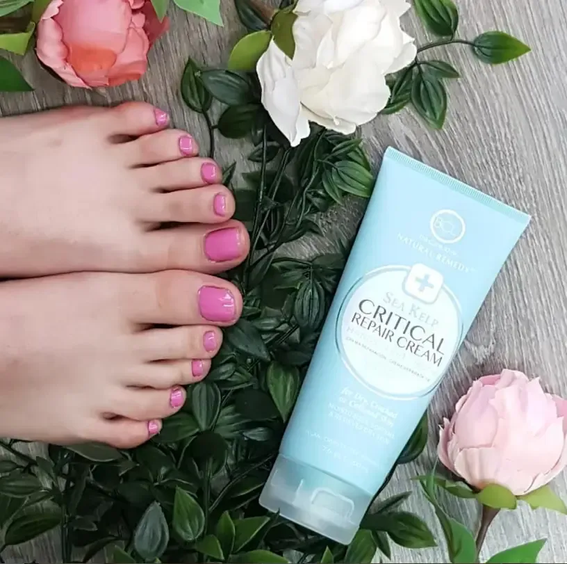 Un tubo de crema reparadora Sea Kiss Critical Repair Cream reposa junto a dos pies con las uñas pintadas de rosa sobre un lecho de hojas verdes.