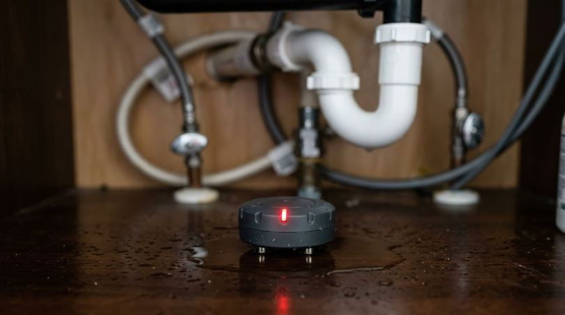 Un détecteur de fuites d'eau circulaire et sombre, muni d'une lumière rougeoyante, posé sur le sol humide.