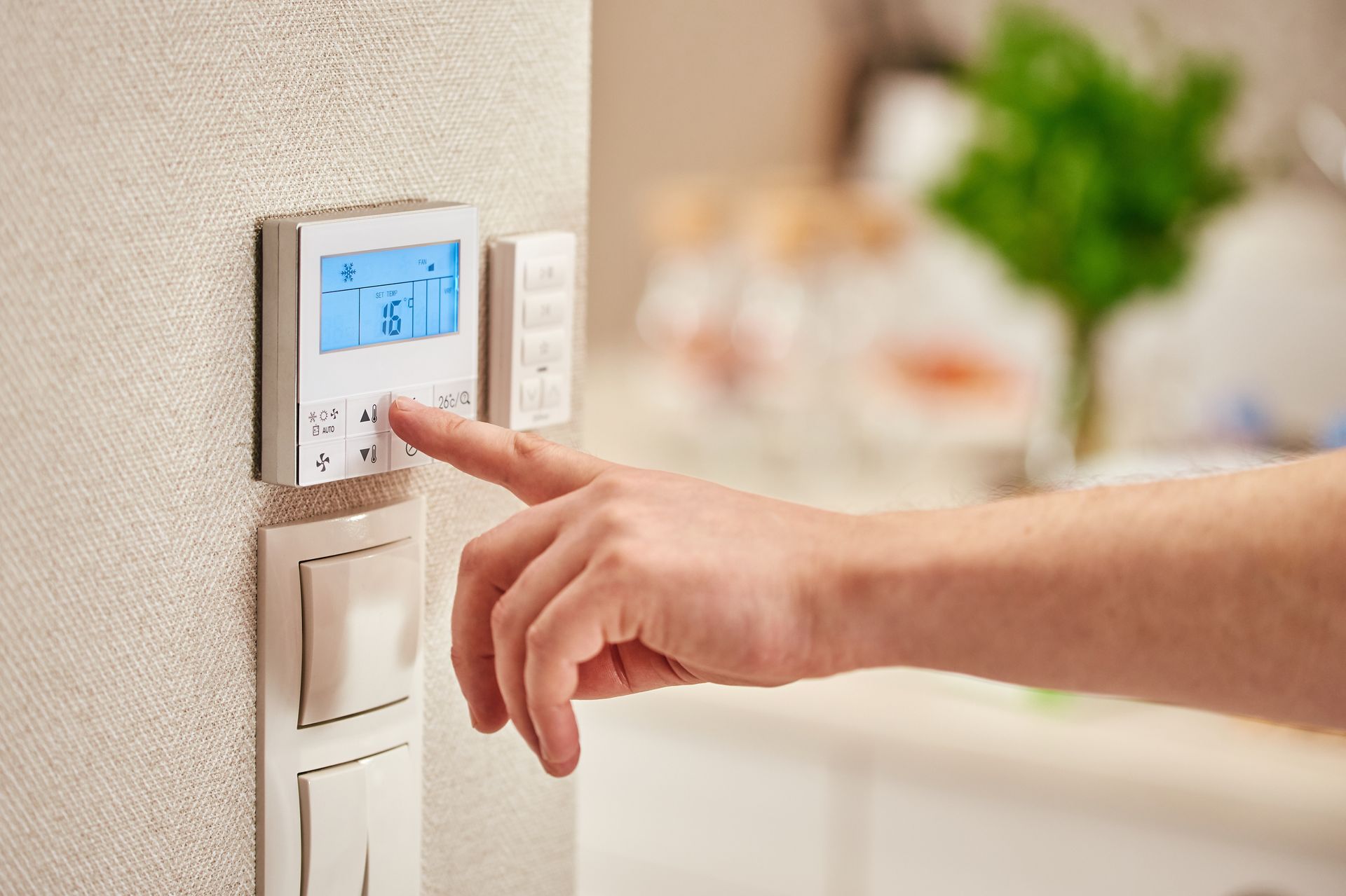 Une main appuie sur un bouton d'un thermostat numérique mural, avec des interrupteurs d'éclairage en dessous, dans une pièce.