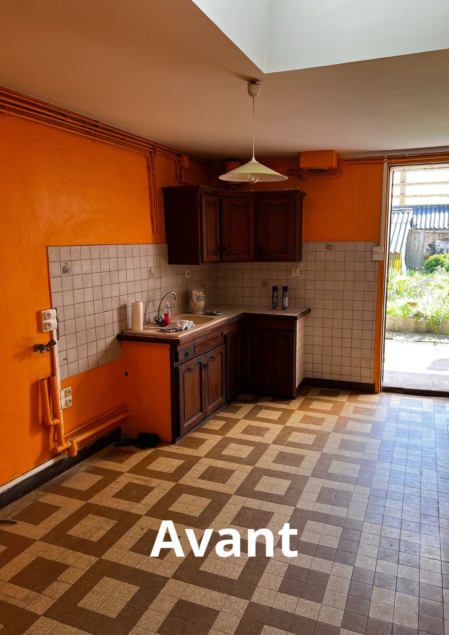 Cuisine aux murs orange, armoires sombres, sol à carreaux et vue sur la cour. « Avant » est écrit en dessous.