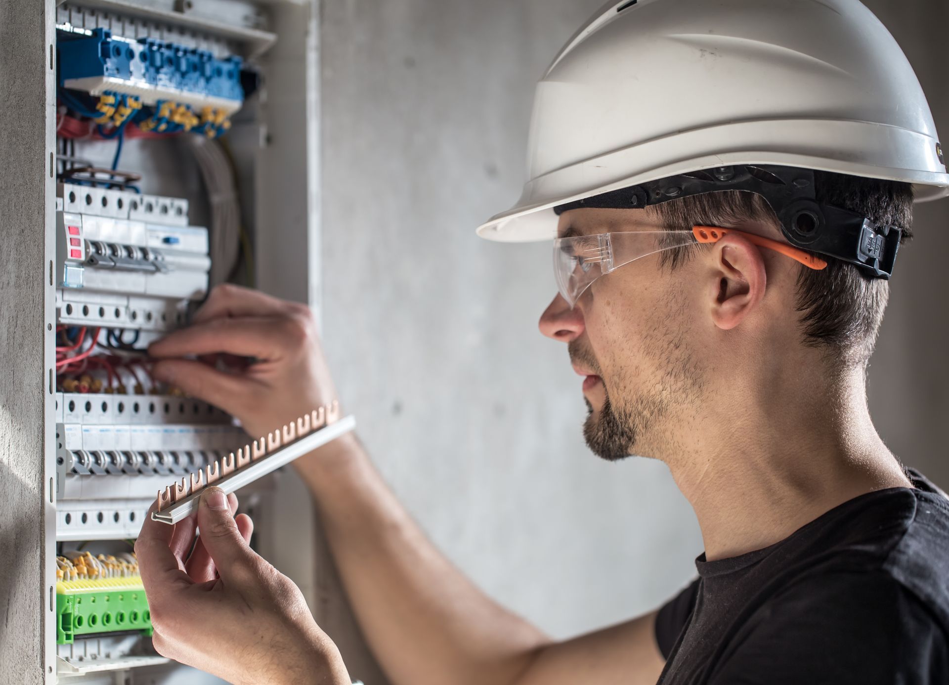 Électricien portant un casque blanc et des lunettes de sécurité travaillant sur un boîtier de disjoncteurs.