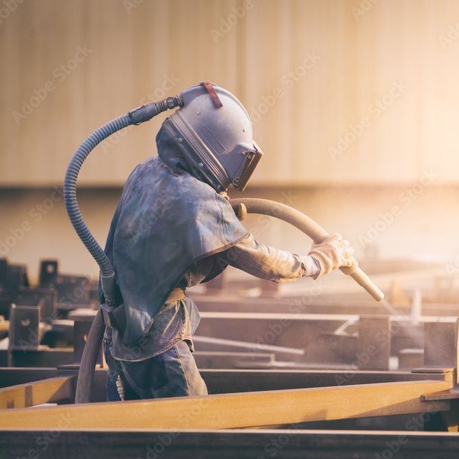 Eine Person mit Helm sandstrahlt ein Stück Metall.