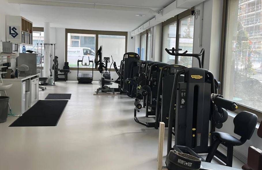Intérieur d'une salle de sport avec des appareils de musculation alignés le long d'une fenêtre. Sols blancs et clairs.
