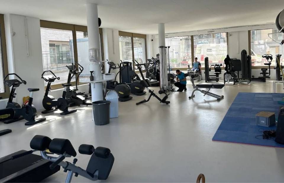 Intérieur d'une salle de sport équipée d'appareils de musculation, notamment des vélos, des tapis de course et des machines de musculation. Plusieurs personnes s'entraînent.