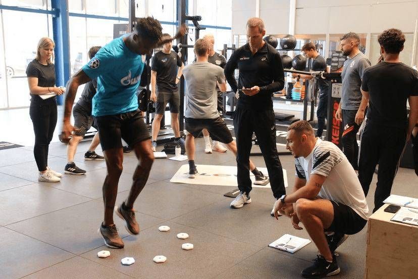 Un homme en chemise bleue saute par-dessus de petits disques, sous le regard des entraîneurs et autres personnes présentes dans une salle de sport.