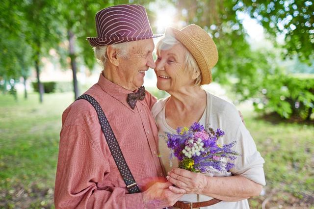 Una pareja de ancianos de pie uno al lado del otro en un parque sosteniendo un ramo de flores.
