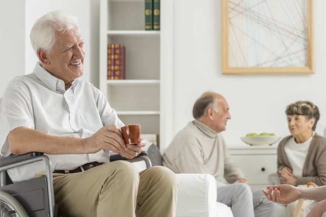Un anciano en silla de ruedas está sentado en una sala de estar sosteniendo una taza de café.