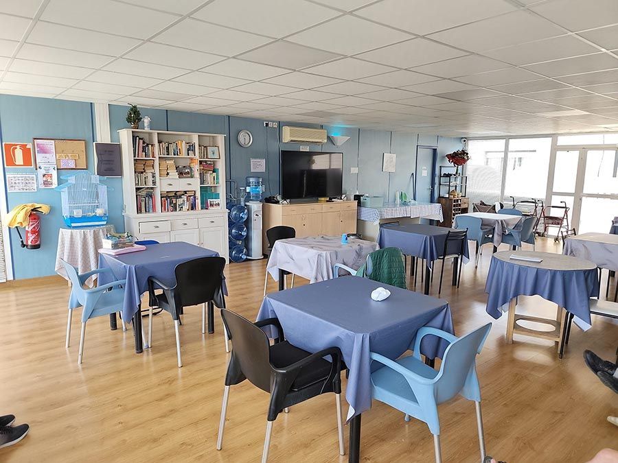 Comedor luminoso con manteles azules, sillas dispersas y grandes ventanales con vistas al agua.