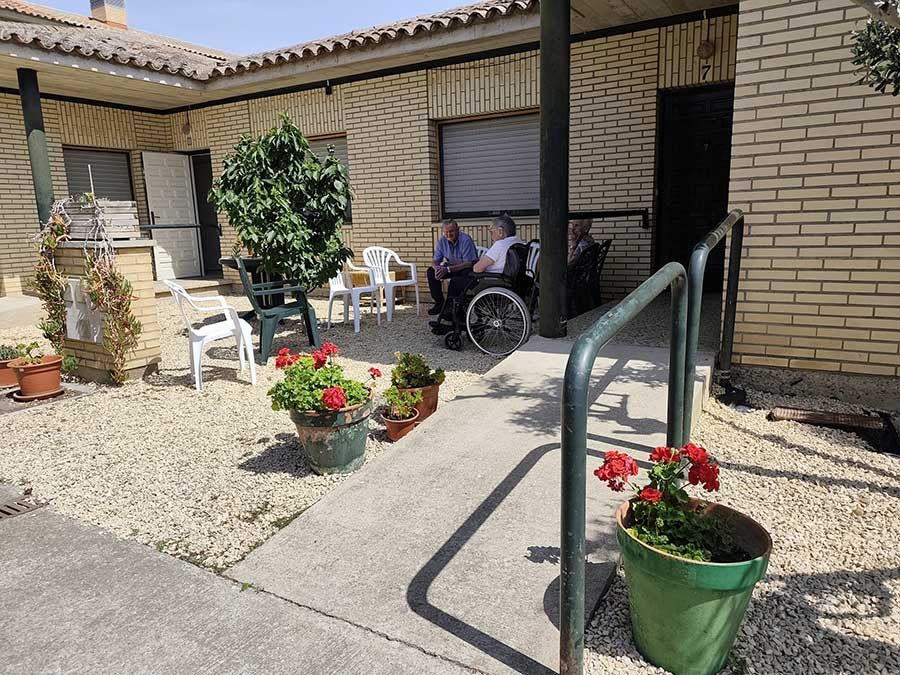 Patio con macetas de flores, sillas y dos personas sentadas en una puerta sombreada.