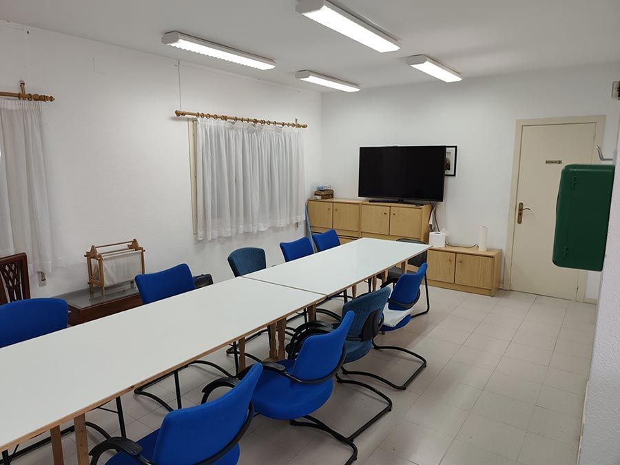 Sala de conferencias con largas mesas blancas, sillas azules, televisor y ventanas con cortinas.