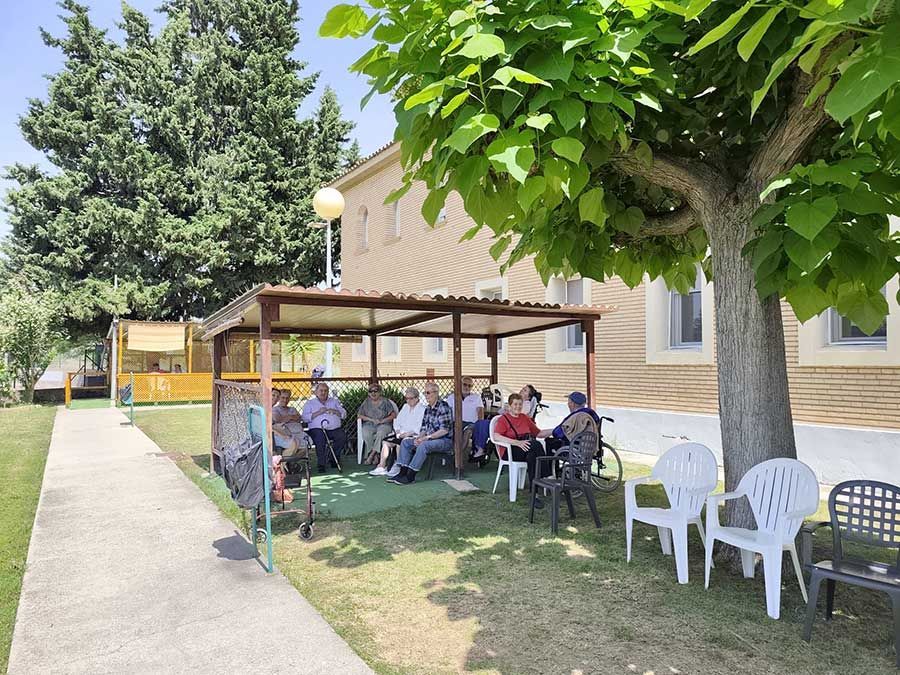 La gente estaba sentada bajo un patio cubierto junto a un edificio de color beige, con sillas y árboles en el jardín.
