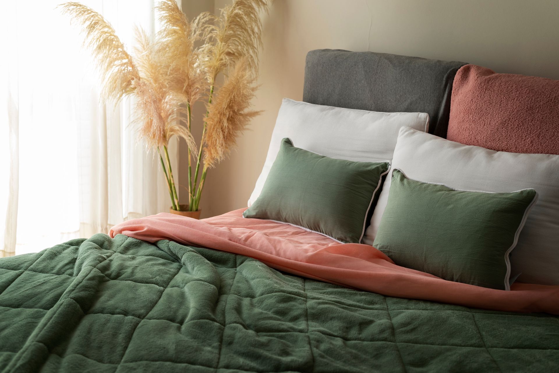 Cama acogedora con edredón verde, manta rosa y almohadas verdes y blancas junto a hierba de la pampa junto a una ventana.