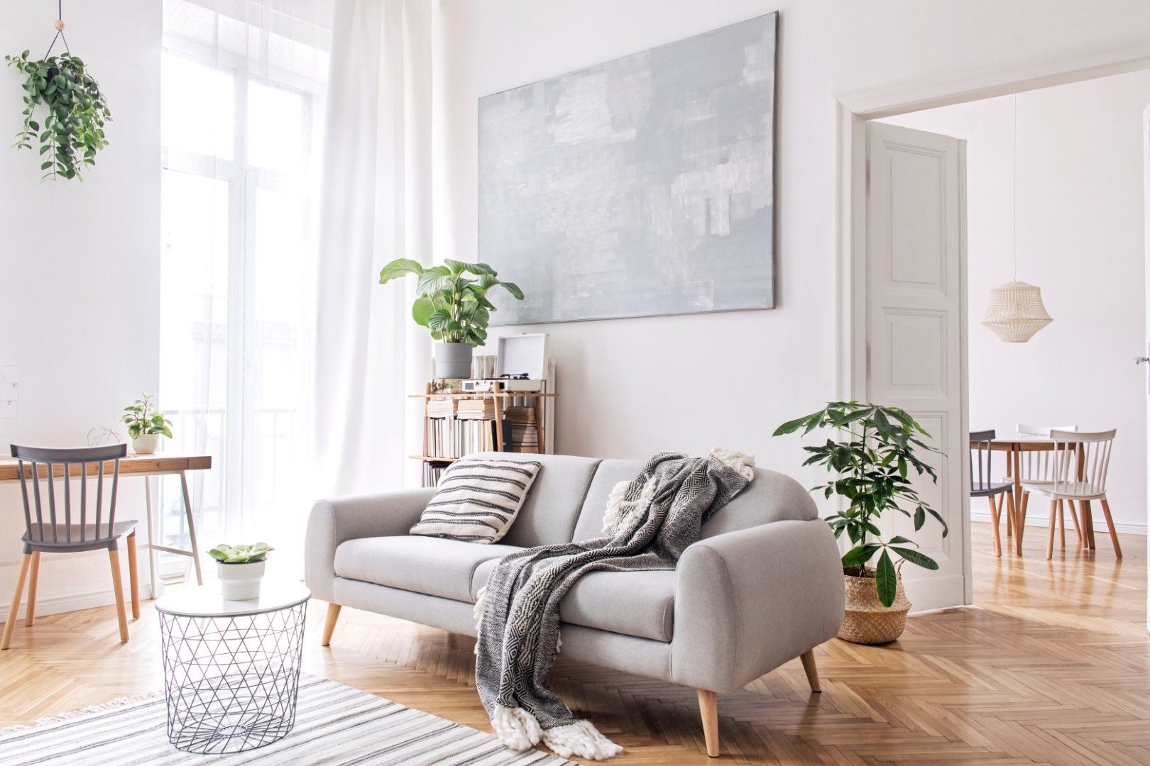 Sala de estar luminosa con un sofá gris, un gran cuadro abstracto en la pared, plantas y suelos de madera.