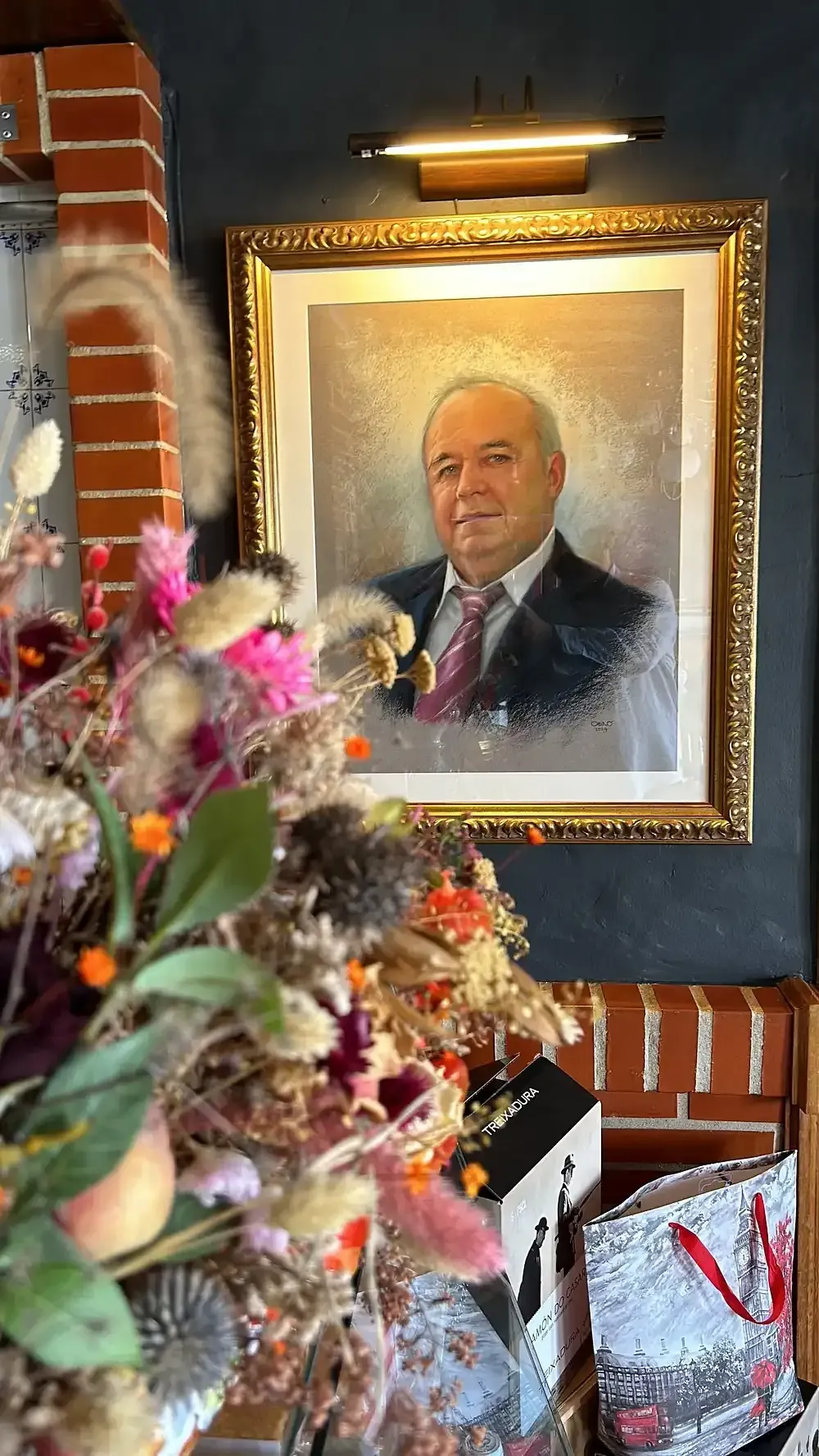 Retrato en marco dorado, iluminado por una pequeña lámpara. Se ve un hombre con traje. En primer plano, un ramo de flores.