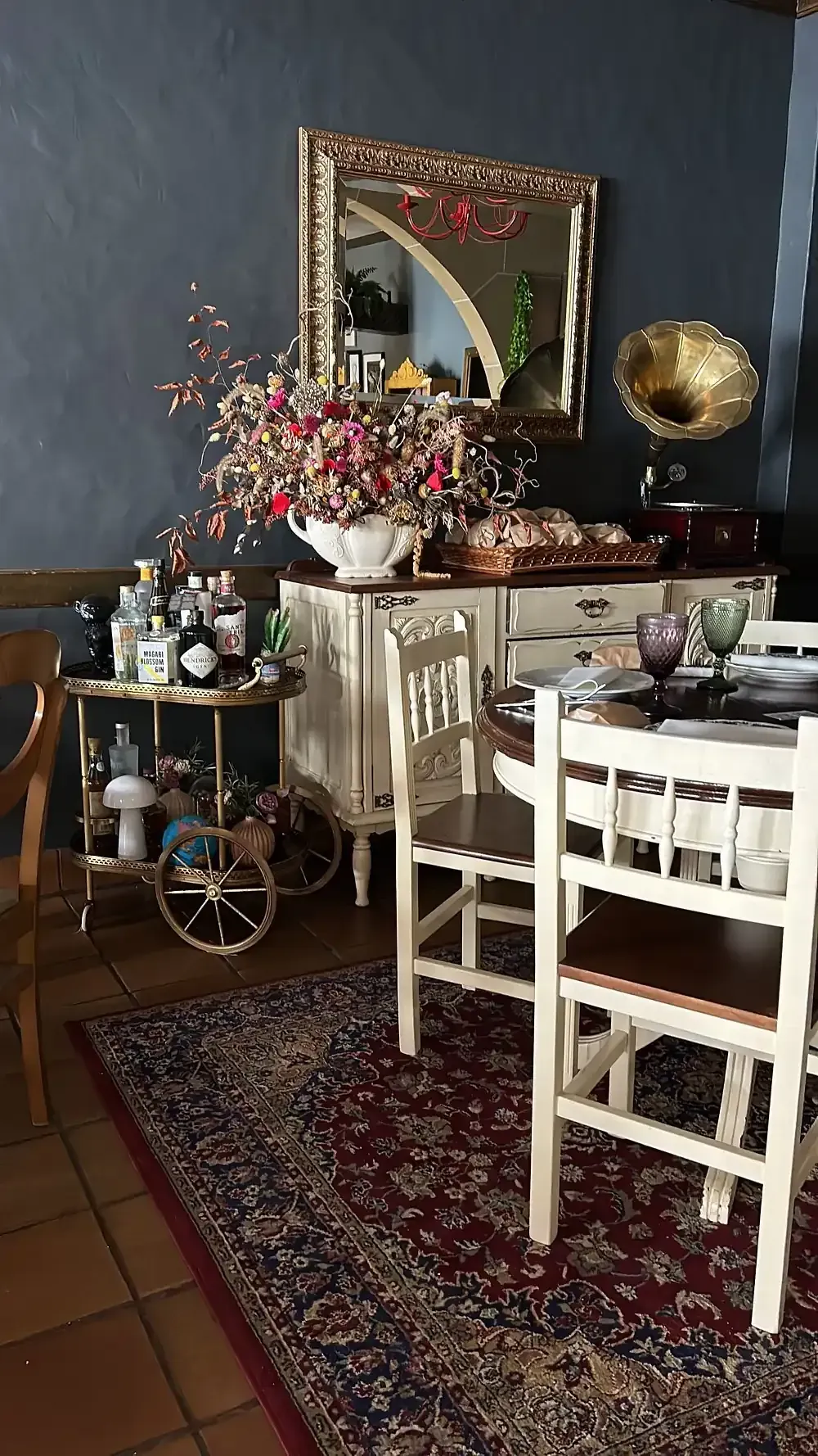 Comedor con mesa de buffet, sillas y carrito de bar. Un espejo con marco dorado cuelga sobre el buffet.