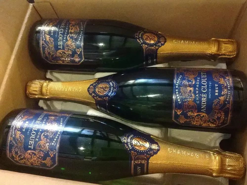 Tres botellas de champán André Clouet en una caja de cartón. Las botellas son verdes, con lámina dorada y etiquetas azules.