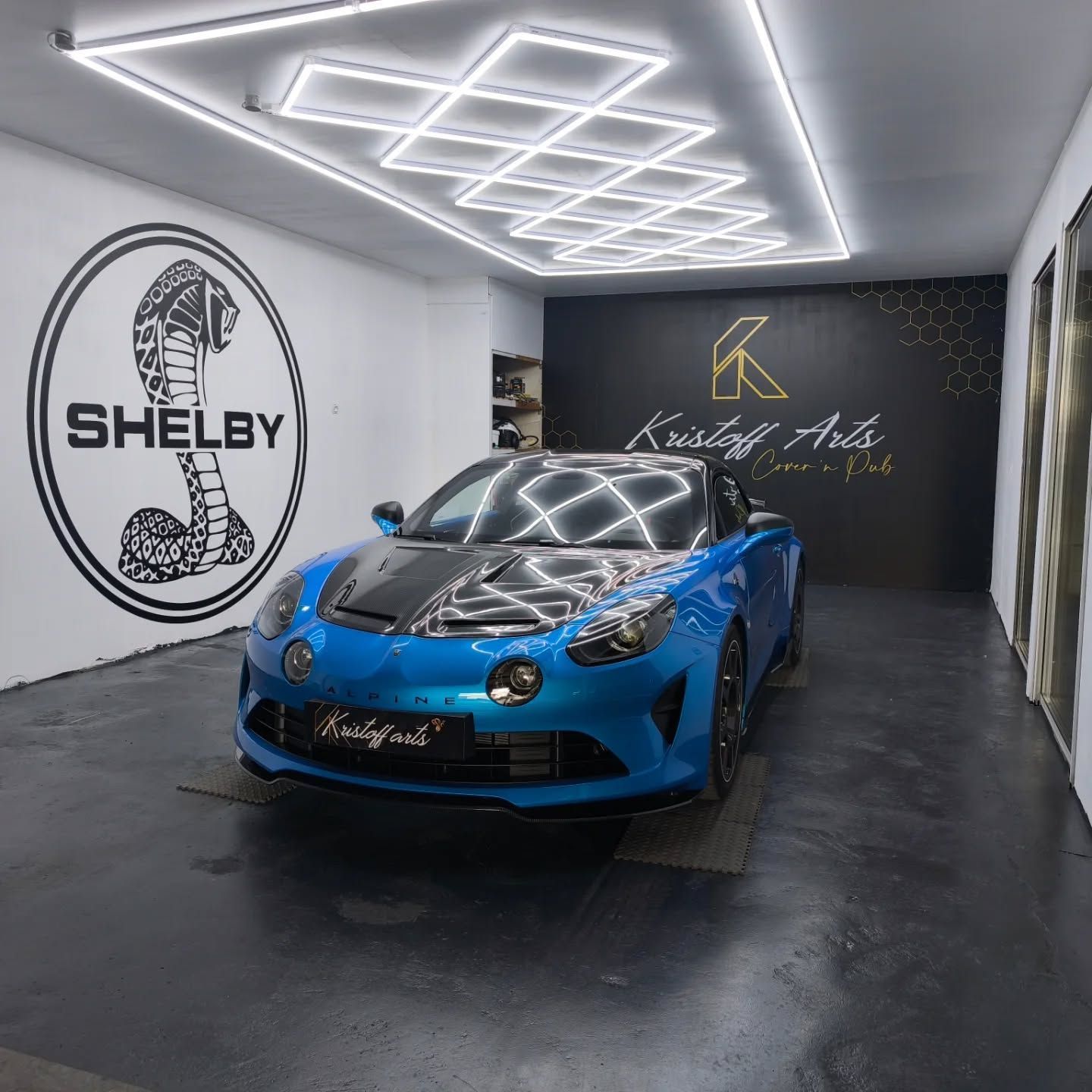 Une voiture de sport Alpine A110 bleue avec un capot en fibre de carbone est garée dans un studio lumineux avec le logo Shelby sur le mur.