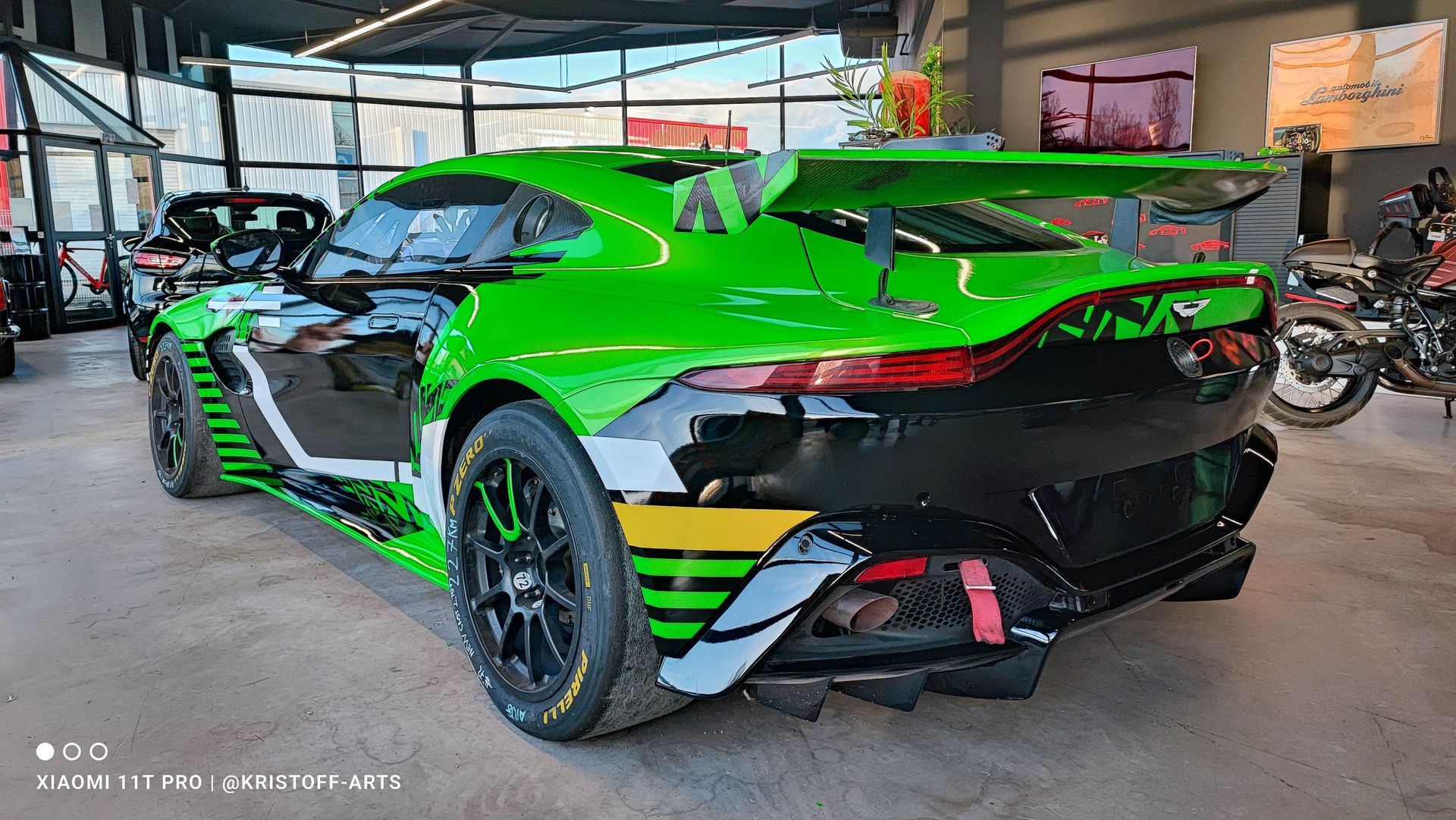 Une Aston Martin GT4 de course vert vif et noir, dotée d'un aileron arrière proéminent, est garée à l'intérieur d'un showroom.