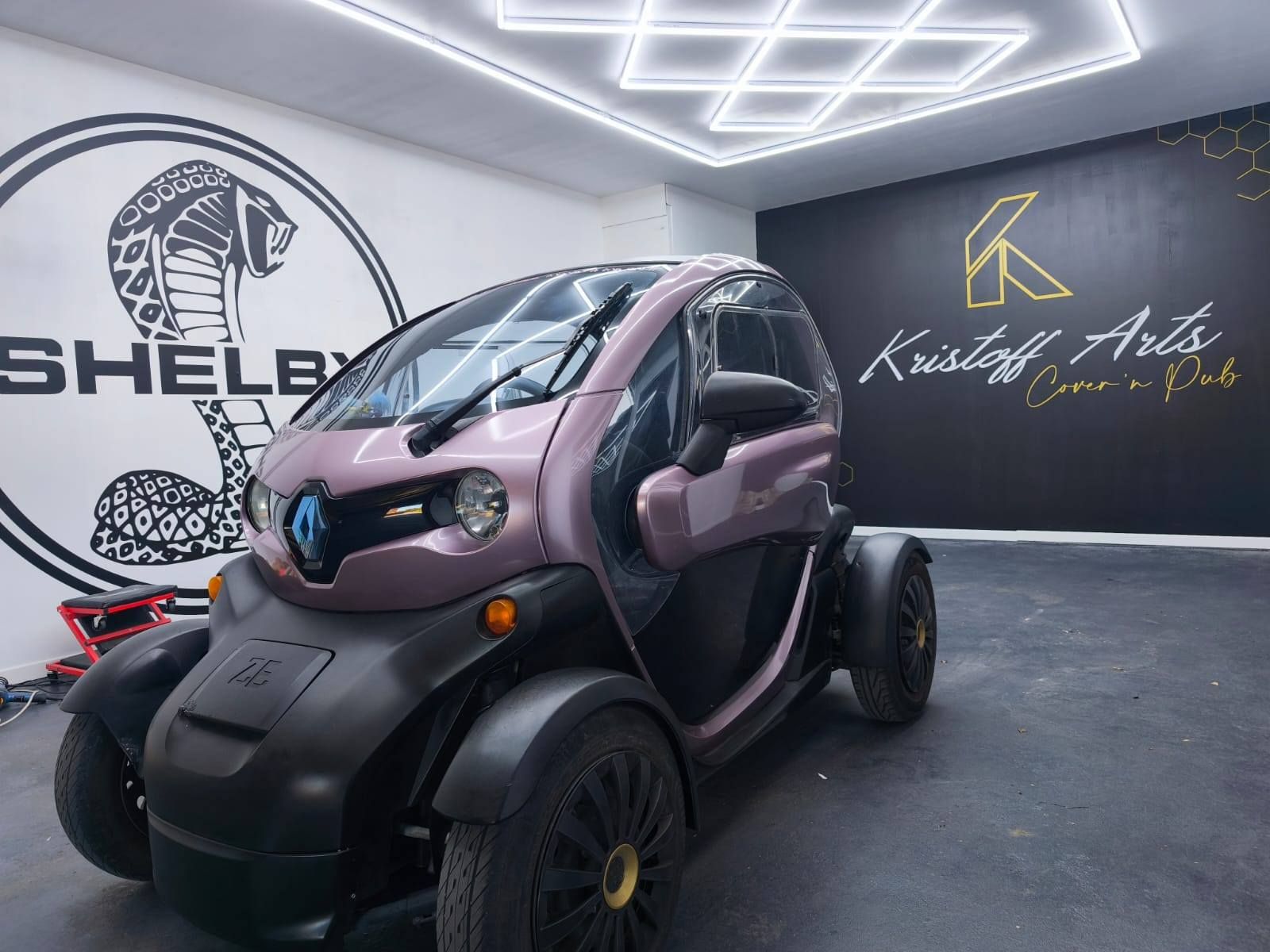 Une petite voiture électrique Renault Twizy mauve garée dans un garage dont les murs sont ornés de grands logos Shelby et Kristoff Arts.