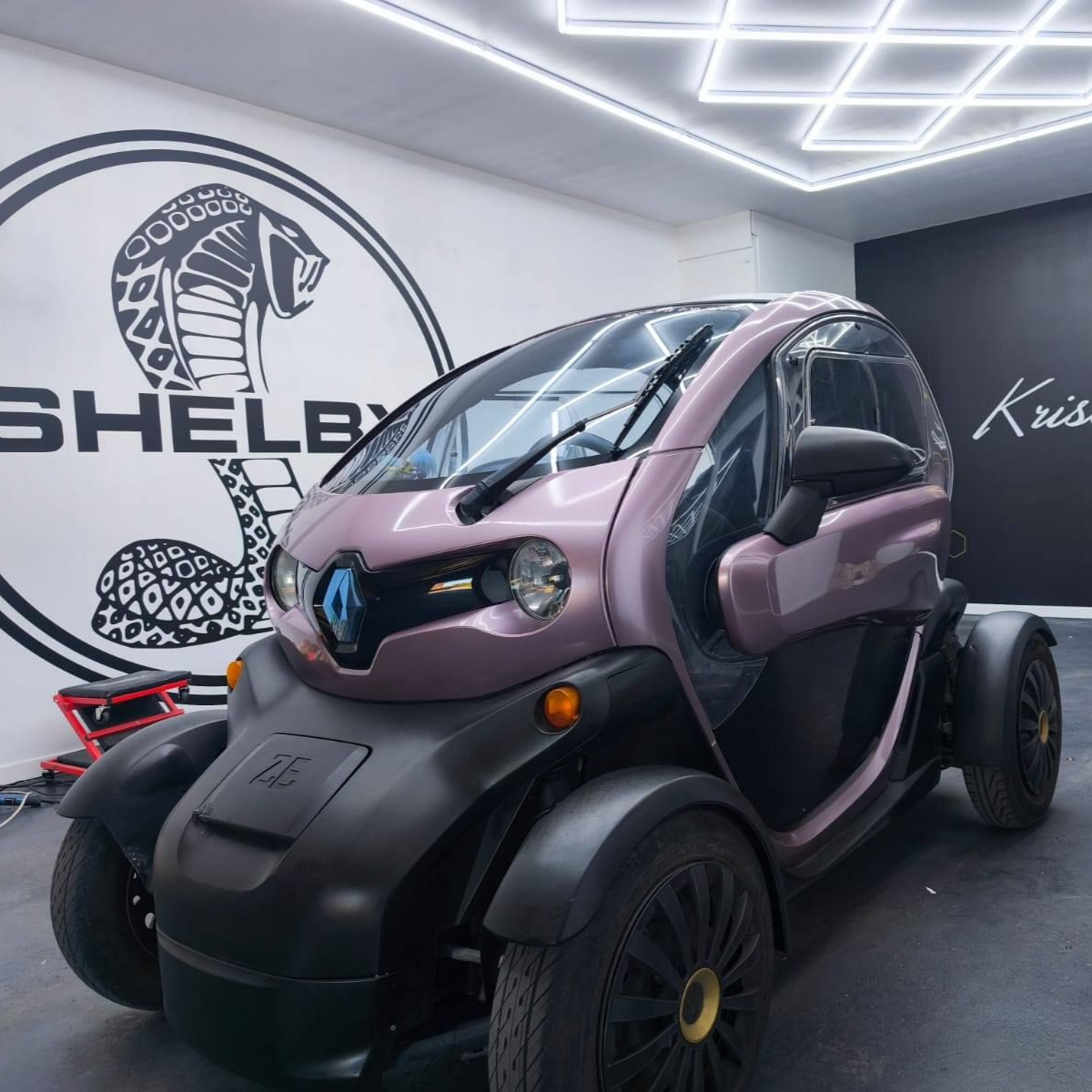 Une petite voiture électrique Renault Twizy violet clair est garée dans un studio, avec un grand logo Shelby Cobra sur le mur derrière elle.