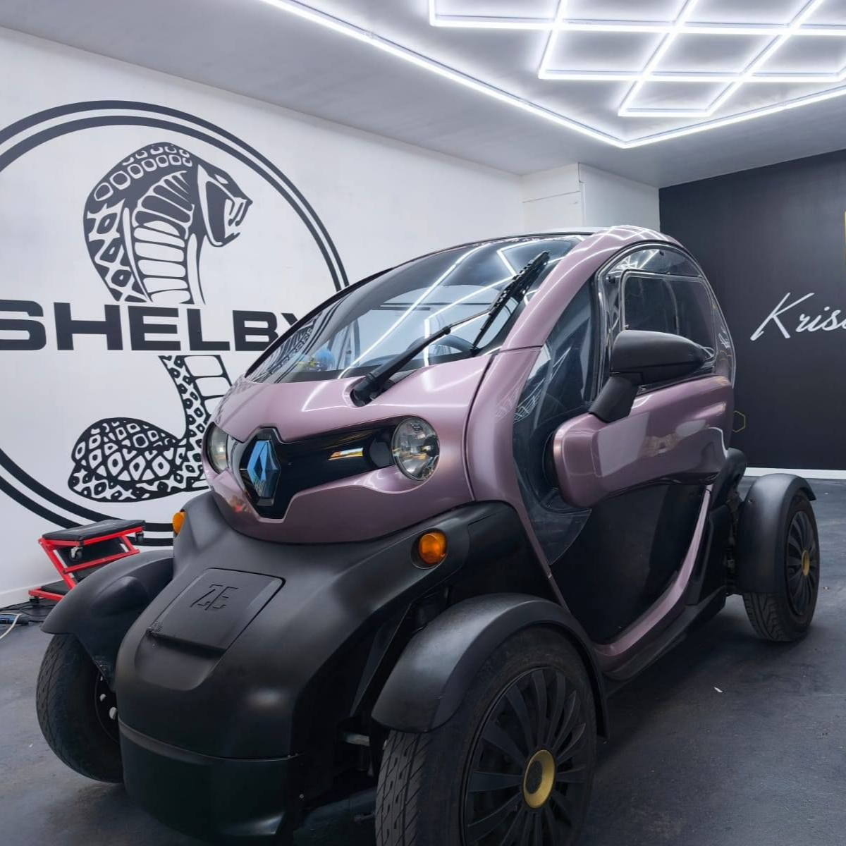 Une Renault Twizy rose garée à l'intérieur, devant un logo Shelby sur un mur blanc.