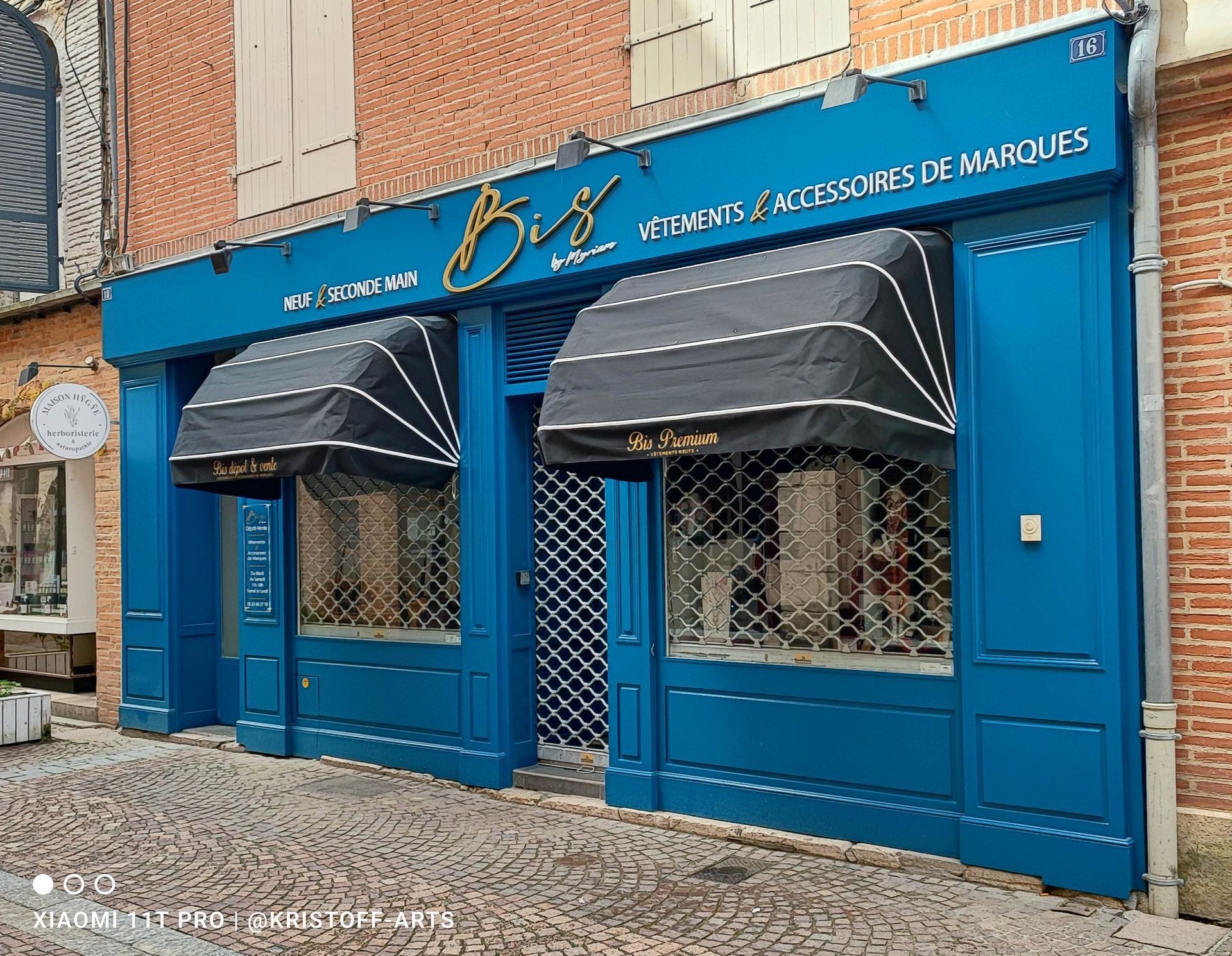 Façade de magasin avec une façade bleue et deux auvents à rayures noires au-dessus des vitrines, munis de grilles de sécurité métalliques.