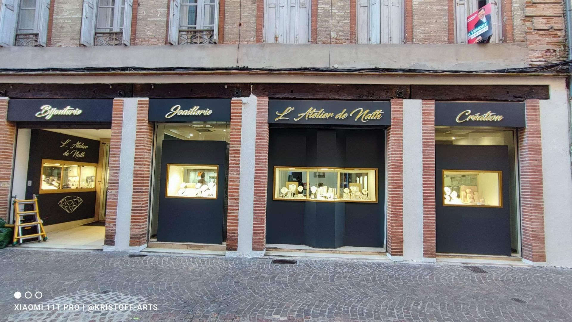 Une devanture en briques comportant trois vitrines à panneaux noirs pour une bijouterie appelée L'Atelier de Nelli.