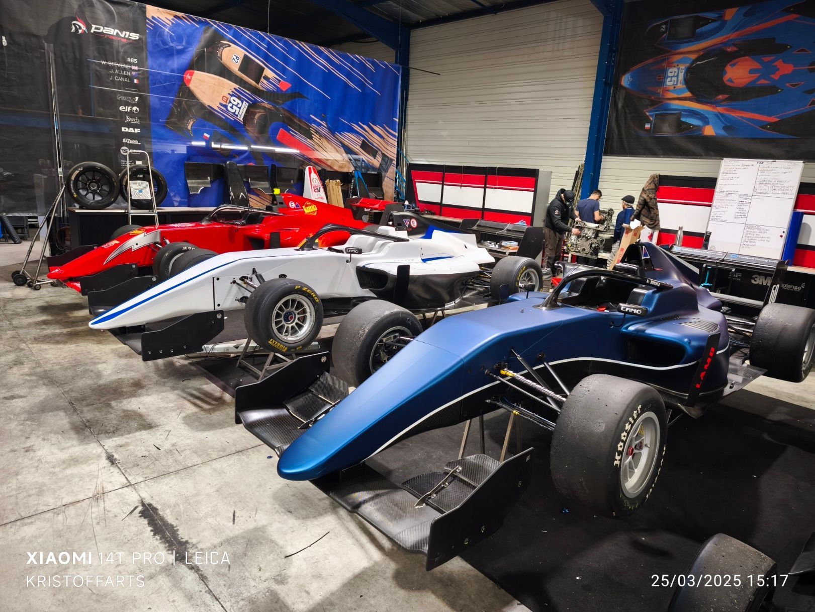 L'intérieur d'un garage expose trois voitures de course monoplaces bleues, blanches et rouges alignées en rang.