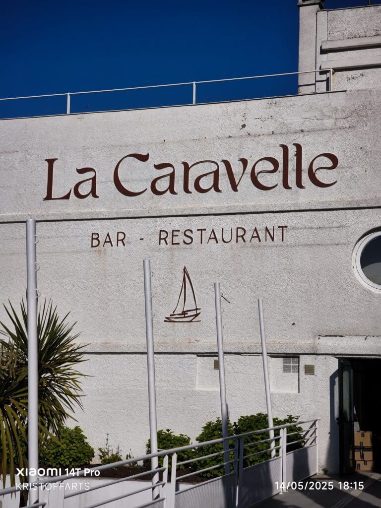 L'extérieur du bar-restaurant La Caravelle, un bâtiment blanc orné d'un logo représentant un voilier et d'une balustrade métallique moderne.