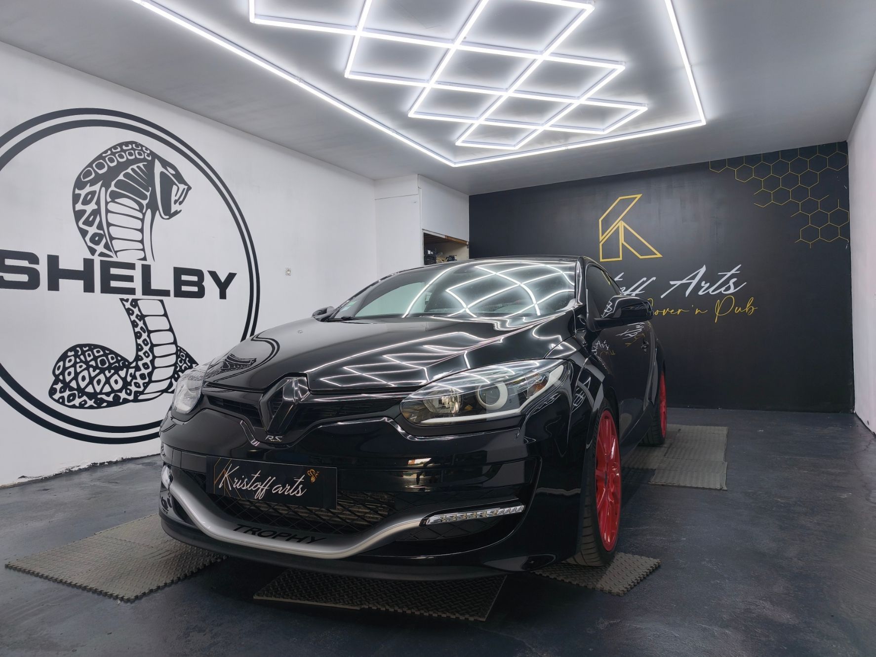 Une Renault Megane noire à jantes rouges est garée dans un studio orné d'un logo Shelby Cobra au mur et de luminaires géométriques au plafond.
