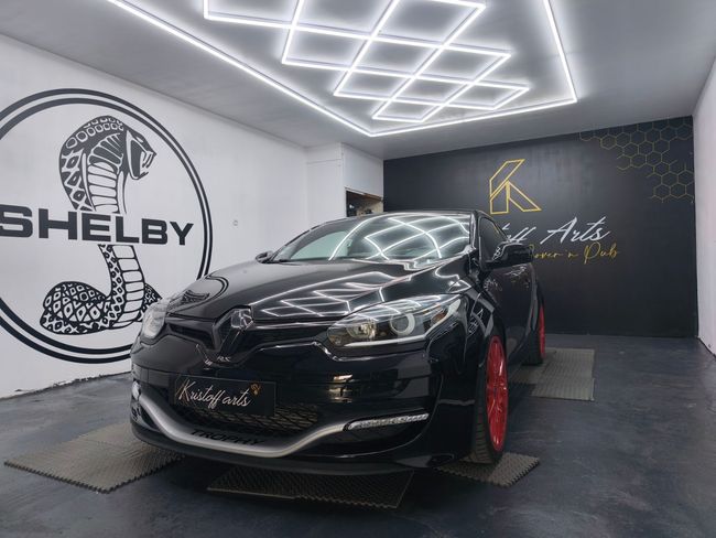 Une Renault Megane noire à jantes rouges trône dans un studio éclairé par des lumières géométriques vives et orné d'un mur arborant le logo Shelby Cobra.