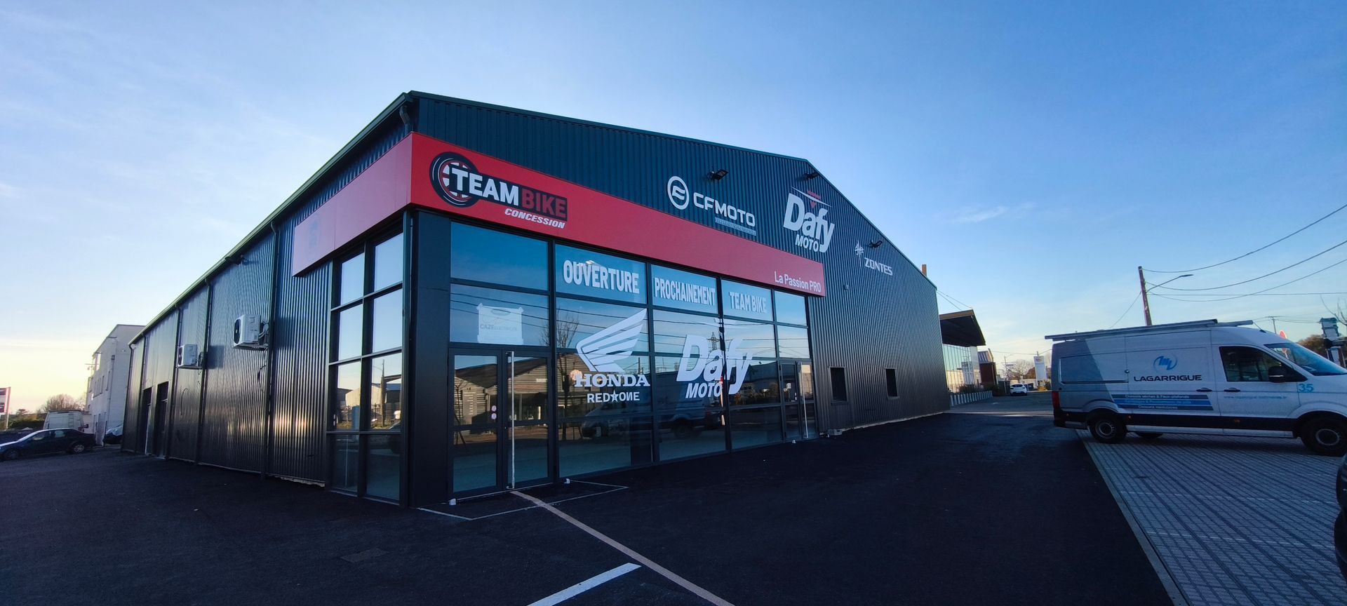 Un bâtiment commercial moderne aux façades sombres, avec des ornements rouges, une devanture en verre et une enseigne Team 500 sous un ciel bleu.
