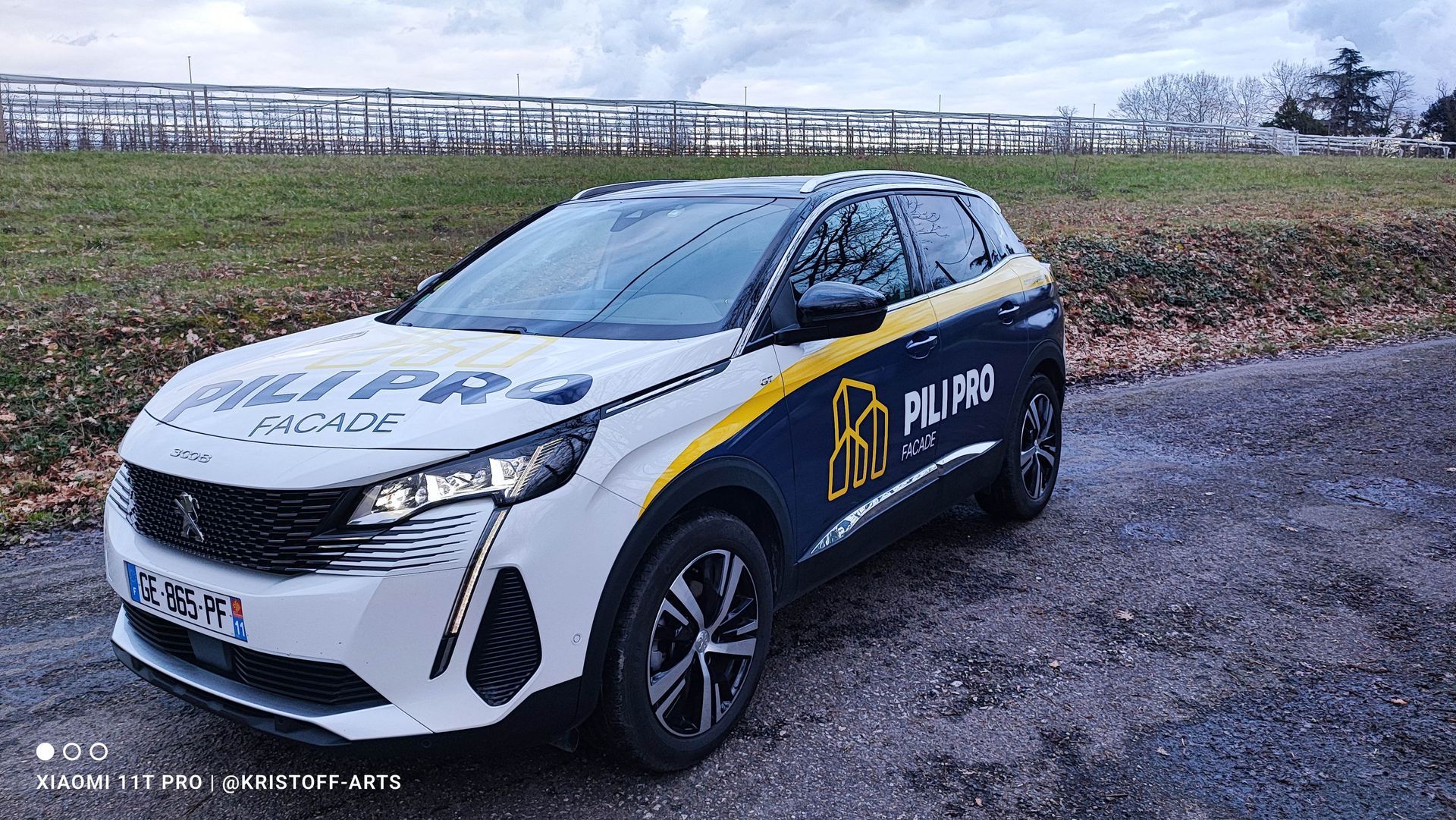 Un SUV Peugeot blanc et bleu foncé, portant l'inscription PILI PRO, est garé sur un chemin de gravier près d'un champ.