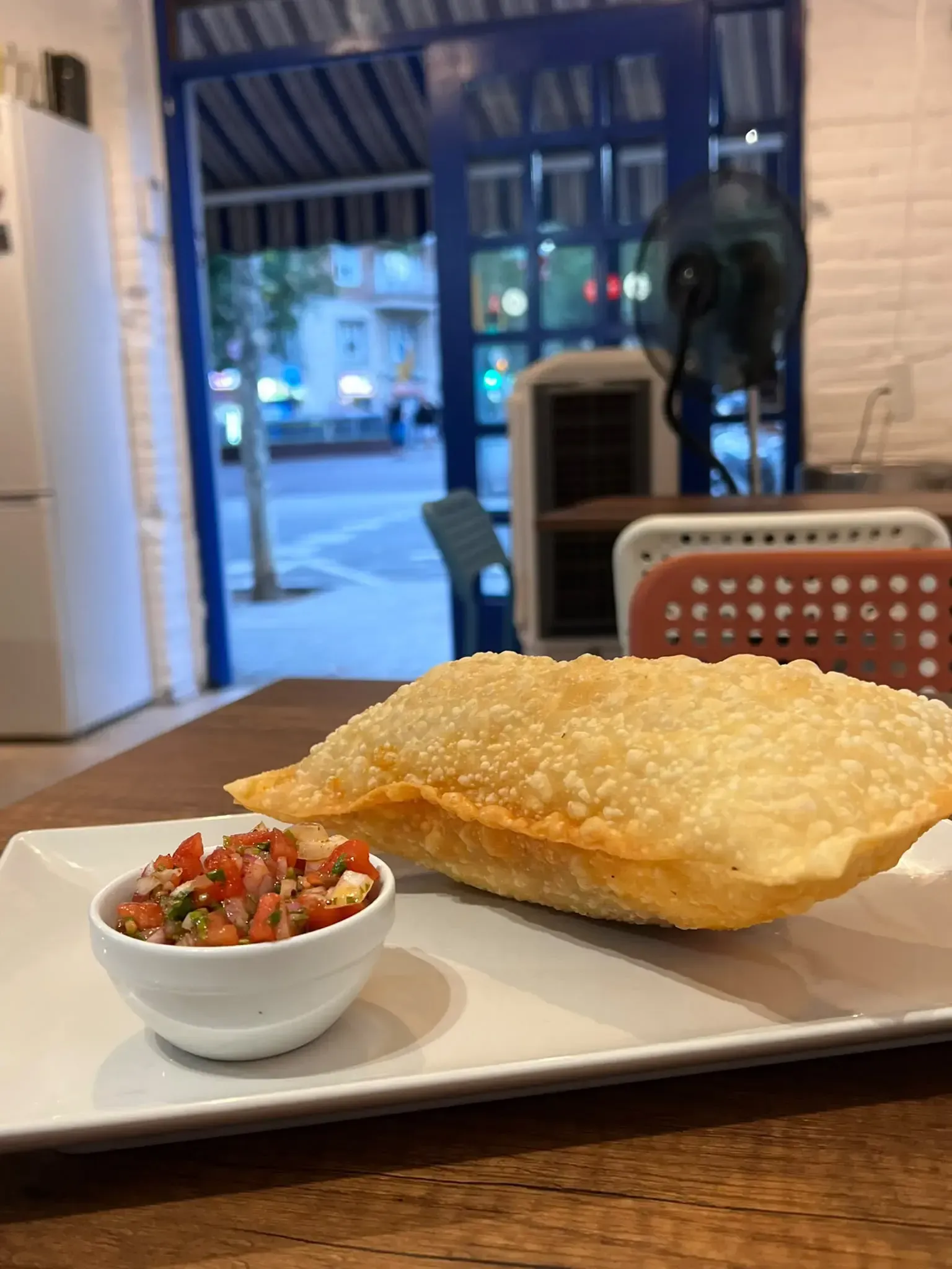 Plato con un pastelito frito y un pequeño recipiente de salsa sobre una mesa de restaurante junto a una ventana.