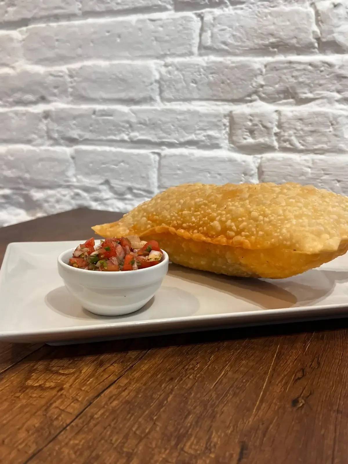 Plato blanco con un pan frito dorado y esponjoso y un pequeño cuenco de salsa sobre una mesa de madera.