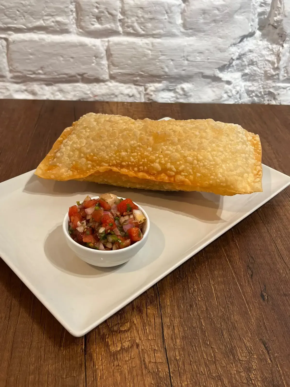 Un pastelito frito dorado en un plato blanco con un pequeño cuenco de salsa roja sobre una mesa de madera.