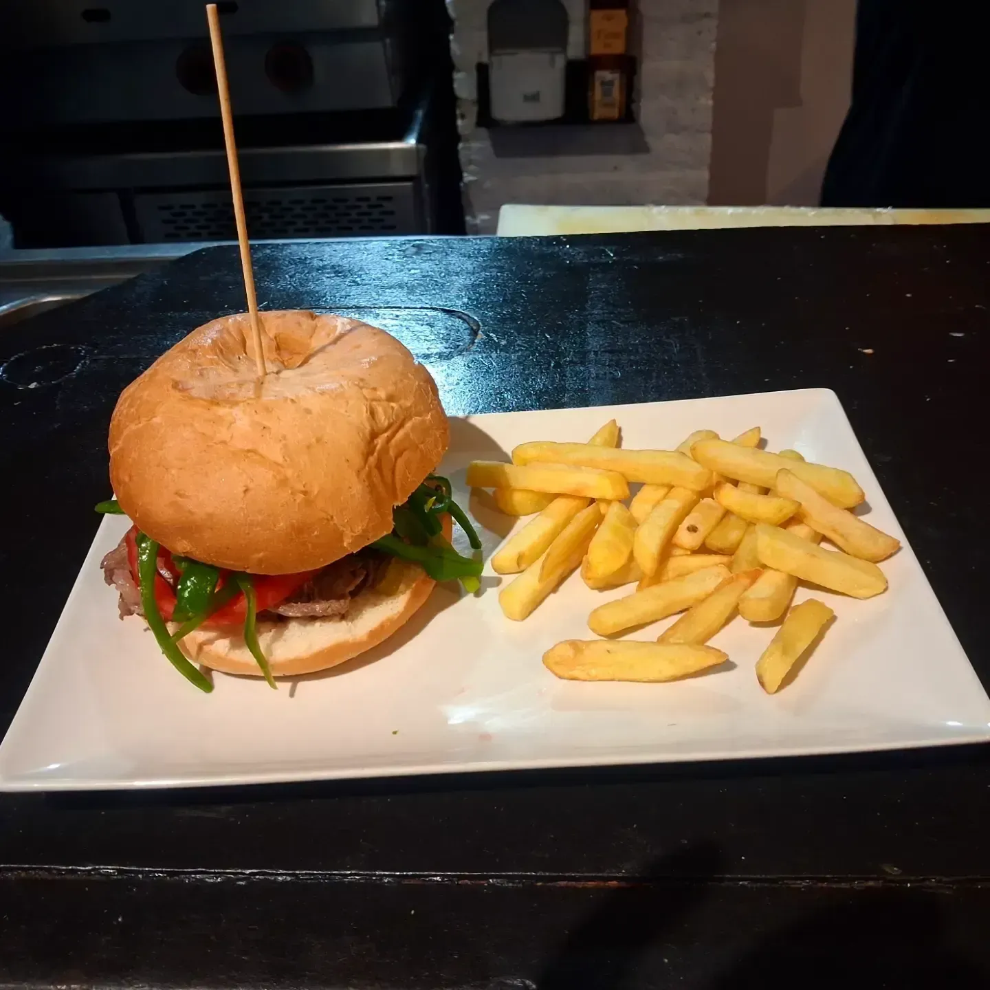 Hamburguesa y patatas fritas en un plato blanco sobre una mesa oscura, con un palillo de dientes clavado en la hamburguesa.