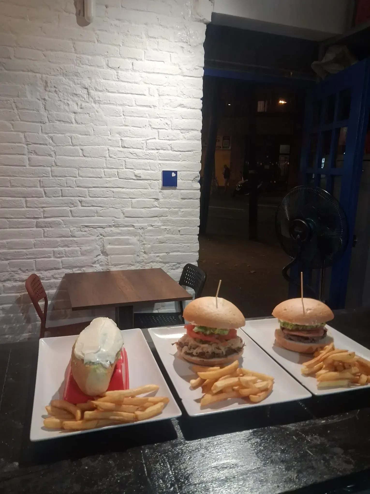 Tres hamburguesas y patatas fritas servidas en un plato sobre una mesa de restaurante, junto a una pared de ladrillo blanco.