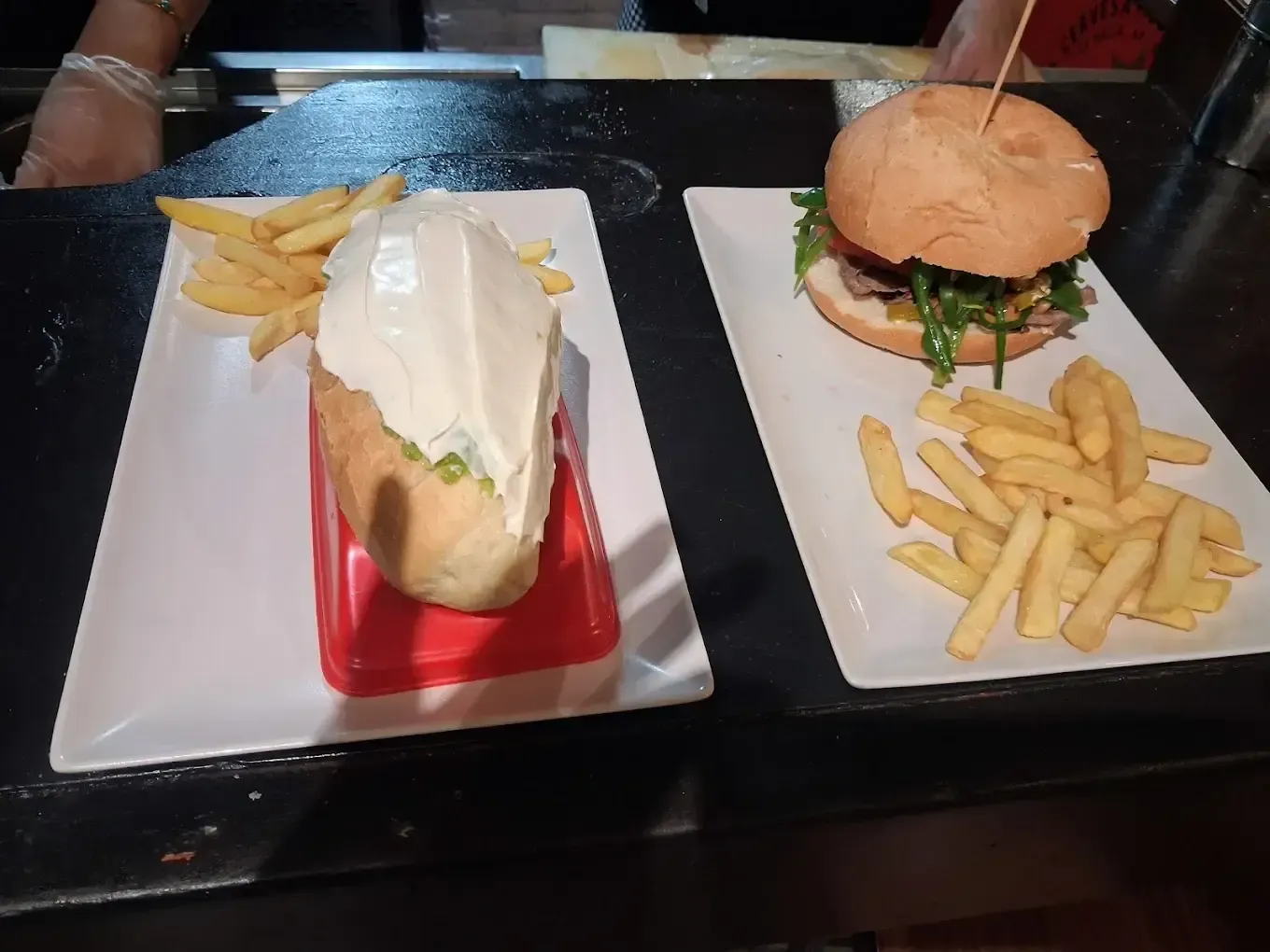 Perrito caliente y hamburguesa con patatas fritas en platos blancos sobre una mesa oscura.