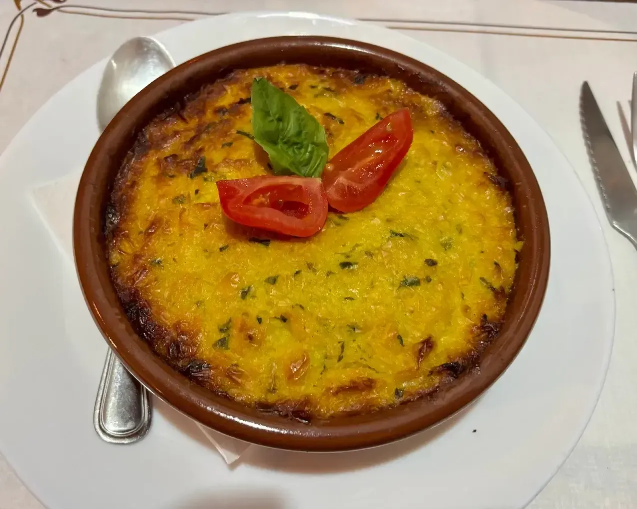 Plato gratinado con queso en un cuenco de barro marrón, adornado con rodajas de tomate sobre un plato blanco.