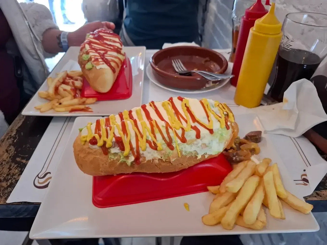 Dos perritos calientes con patatas fritas, kétchup y mostaza sobre una mesa en un restaurante.