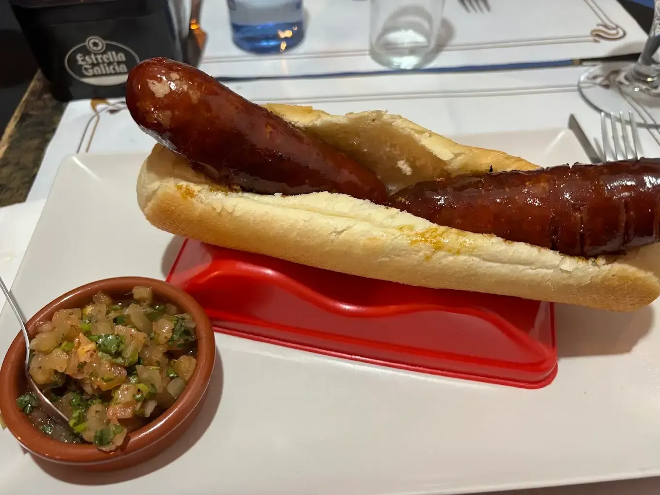 Salchicha a la parrilla en un panecillo sobre un plato blanco con una guarnición de salsa en una mesa de restaurante.