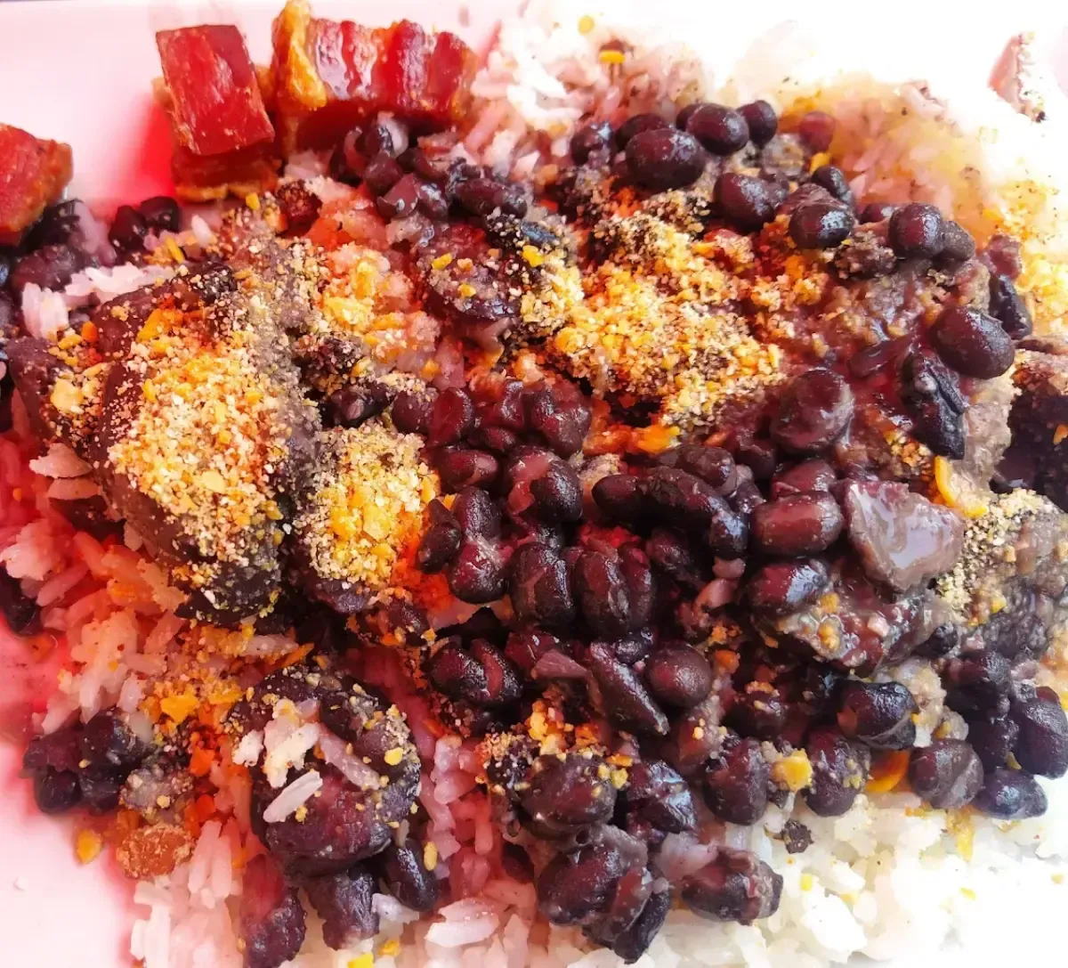 Frijoles negros cubiertos con pan rallado, acompañados de plátanos fritos y arroz en un plato rosa.