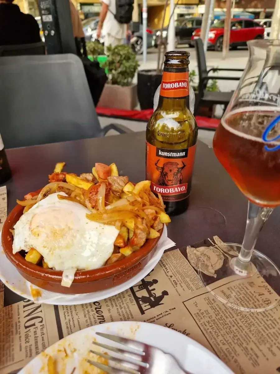 Un plato de huevos fritos y patatas sobre la mesa de un café, junto a una botella de cerveza y un vaso de bebida ámbar.