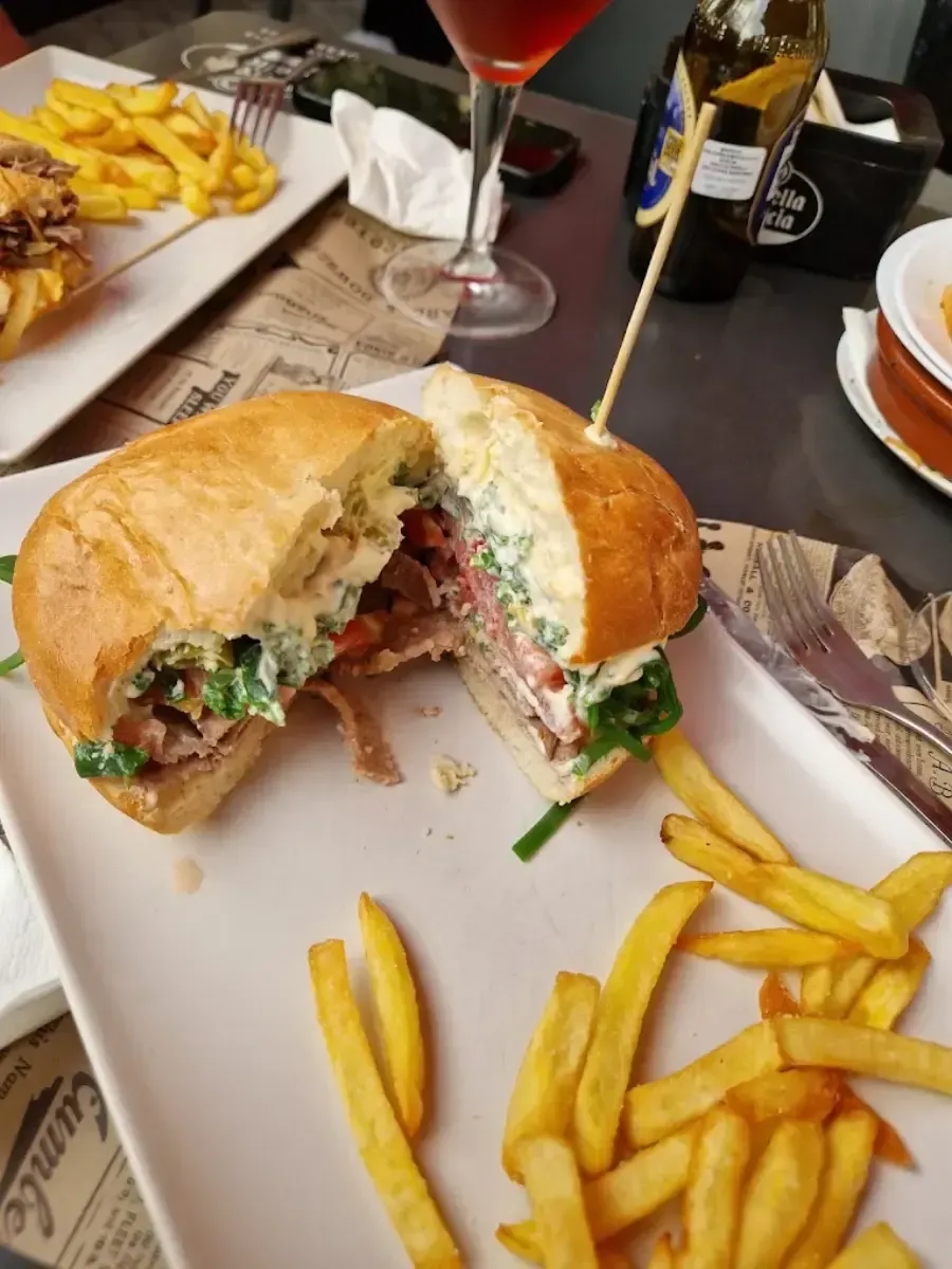 Sándwich cortado con patatas fritas sobre la mesa de un restaurante, con un cóctel rojo al fondo.