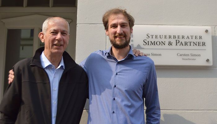 Zwei Männer posieren für ein Foto vor einem Schild mit der Aufschrift „Simon & Partner“