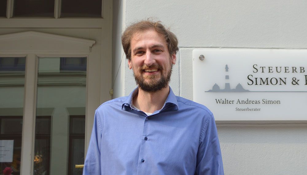 Ein Mann in einem blauen Hemd steht vor einem Schild mit der Aufschrift „Steuerb Simon & I“.